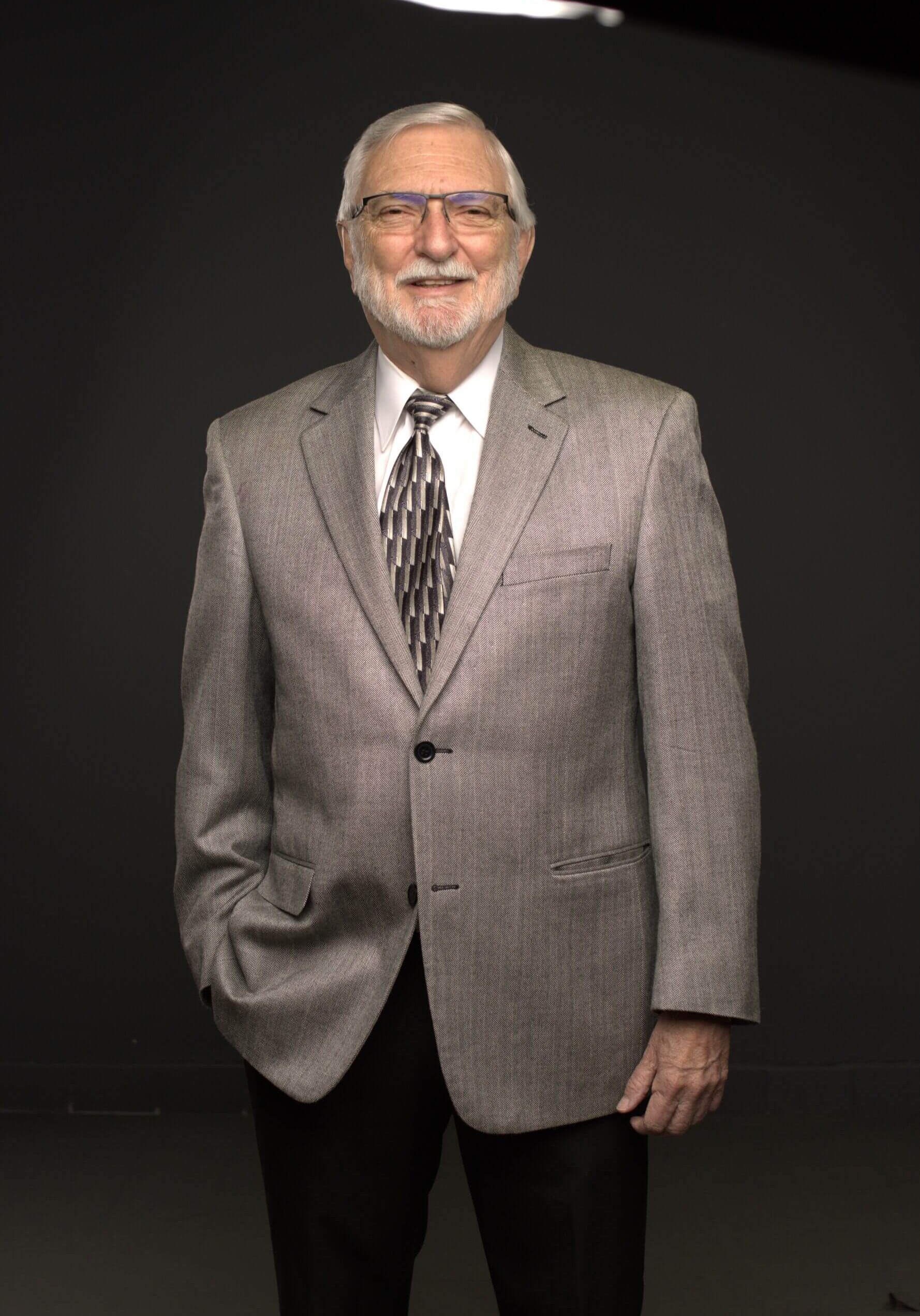 Elderly man in a gray suit and tie with hands in pockets.